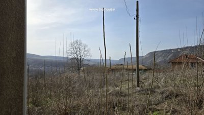 View over dry plants and rooftops towards rolling hills in the distance under a clear sky