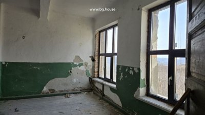 Interior room showing two large windows, peeling white and green walls, and wooden floor