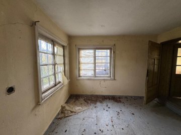 An empty room with two windows, each partially covered with torn paper, and a wooden floor scattered with debris.