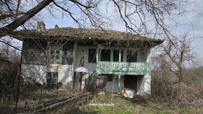 Exterior view of an old rural house featuring a tiled roof and a green wooden balcony with surrounding leafless trees and wire fence.