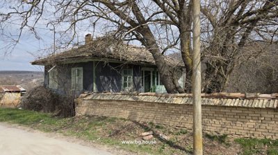 Old rural house showing green window frames and a wooden balcony behind a brick wall with leafless trees in front.