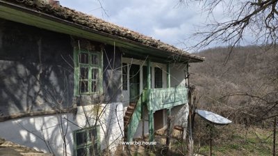 Side view of an old rural house with green windows and wooden balcony behind a brick wall and leafless trees.