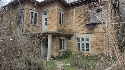 Front corner view of an old two-story brick house with white framed windows and doors, surrounded by dry bushes and grass.