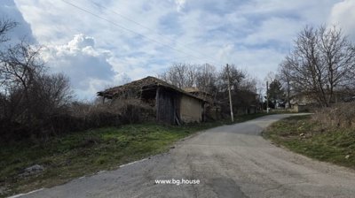 A paved rural road curves past old wooden and stone outbuildings with leafless trees and cloudy sky above.