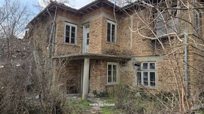 Close-up of the front corner of an old two-story brick house showing white framed windows and doors with surrounding dry vegetation.