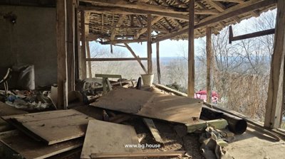 Inside view of a wooden structure with exposed beams, scattered wooden boards, and debris on the floor, overlooking trees outside.