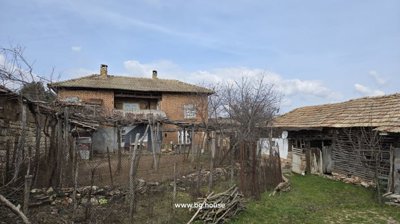 View of a two-story brick house with a tiled roof, wooden outbuildings, and a fenced garden area with leafless trees and vines