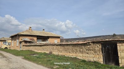 Brick house enclosed by a stone garden wall, with green grass along the adjacent dirt road under a partly cloudy sky
