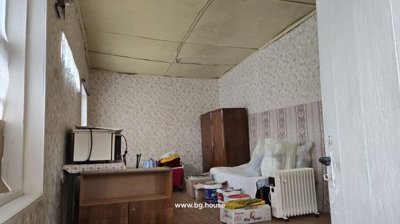 Room interior featuring wallpapered walls, a wooden wardrobe, a bed with pillows, and various household items scattered around
