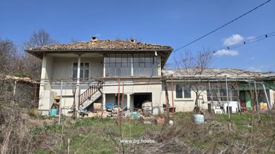 Front facade of a two-storey Bulgarian house showing external stairs and storage area beneath, with dry grass in the foreground