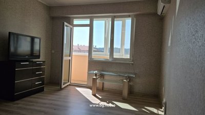 Spacious living room showing a TV on a black chest of drawers, a glass coffee table, and an open door to the balcony.