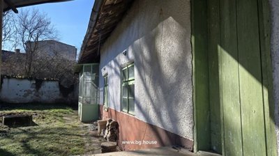 Close-up view of a white plastered rural house wall showing green doors and windows, with garden area featuring grass and scattered roof tiles.