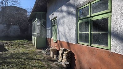 Side view of a white plastered rural house with green doors and windows, adjacent to a garden area with grass and wood piles.