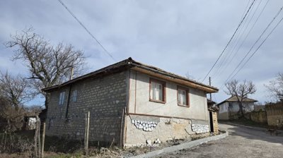 Side and front corner of a Bulgarian village house with a tiled roof, two windows, and a fenced yard beside a dirt road.