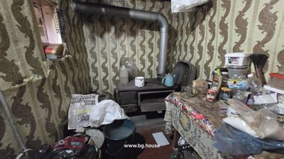 Interior of a small room showing a wood stove with a metal pipe, patterned wallpaper, and a table cluttered with various items.