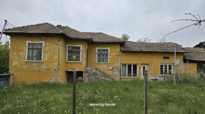 Front view of a yellow rural house with a tiled roof and stone steps leading to the entrance, enclosed by a wire fence with green grass and dandelions