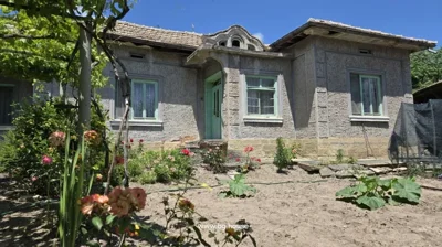 Close-up of the front entrance of a rural house featuring a green door and windows, with flowers and garden plants in the foreground.