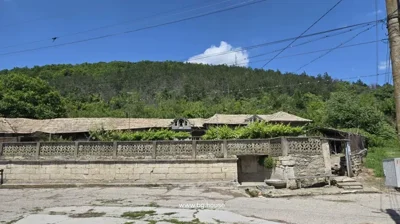 View of a rural house behind a decorative stone fence, with pine forest covering the hillside behind the property.