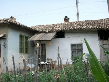 Exterior view of a traditional Bulgarian village house with a tiled roof and garden plants enclosed by a rustic wooden stick fence.
