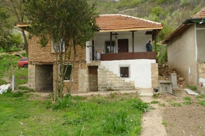 Front view of a two-storey rural Bulgarian house featuring a wooden porch, stone stairs, and a tiled roof.