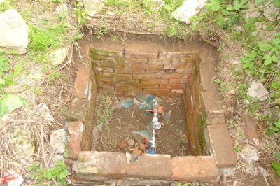 Close-up of a brick-lined pit containing a water valve, surrounded by grass and stones.