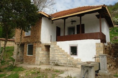Front and side angle of the rural house highlighting stone stairs leading to a wooden porch and tiled roof.