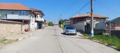 View along a village street showing houses on both sides and cars parked along the roadside under a clear sky.