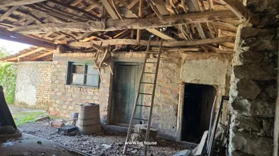 View of a rustic interior showing wooden beams, a ladder leaning on a brick wall with a door and window, and various items including a barrel and a chair hanging from the ceiling