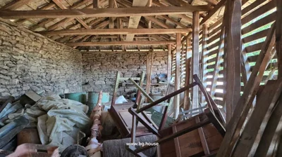 Interior view of a rustic storage area with stone walls, wooden beams, and various stored items such as barrels and wooden furniture