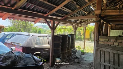 Covered outdoor area supported by wooden posts with barrels lined up and parked cars visible outside in a garden setting