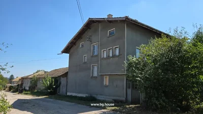 A large three-storey house with several windows and a sloped roof, situated next to a dirt road with greenery around.