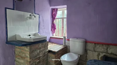 Bathroom corner showing toilet, sink, window with curtain, and wooden cover on brick structure.