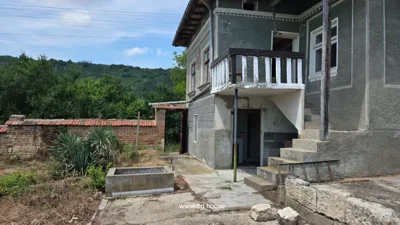 Two-story house with stairs, balcony, brick and stone fence, concrete path, and plants in yard.