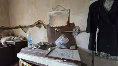 A room showing a sink with a water tap, a mirror above it, a coat hanging on the wall, and various items including a water bottle and framed picture on the sink.