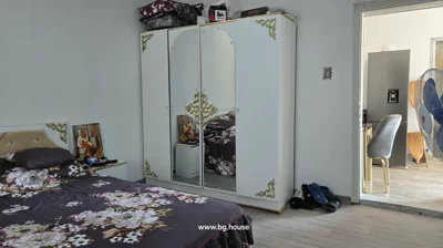 Room with bed covered in floral bedding, white wardrobe with gold decorations and mirrors, and open door revealing kitchen area with refrigerator and fan