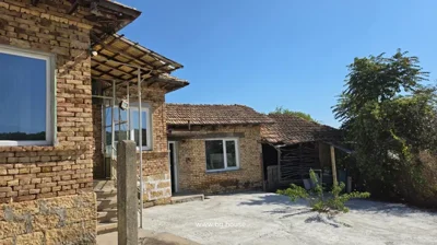Brick house with tiled roof, white-framed windows, concrete yard, and some vegetation under a clear blue sky.