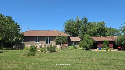 A single-story house with a tiled roof and brown walls, surrounded by a green lawn, trees, and garden beds under a clear blue sky.