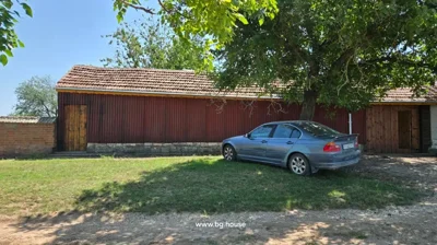 A blue car is parked on grass beside a long building with red corrugated metal siding and wooden doors, shaded by trees.