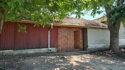 A building with red corrugated metal siding and wooden double doors stands next to a tree casting shadow on the dirt ground.