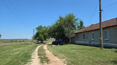 A dirt road runs alongside grass, trees, two parked cars, and a long building with multiple windows under a clear sky.