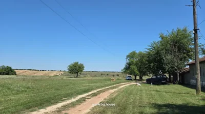 A dirt road bordered by grass and trees leads past two parked cars near a building, with open fields visible in the background.