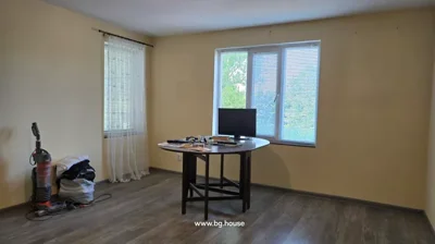 Room featuring two windows with blinds and sheer curtains, a round table with a monitor and miscellaneous items, and a vacuum cleaner with bags on the floor