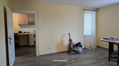 Room showing an open door to a kitchen area, vacuum cleaner and bags on the floor, and a window with sheer curtain
