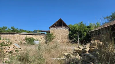 Exterior view of stone building with tiled roof and wooden upper section, surrounded by dry grass and stone piles.
