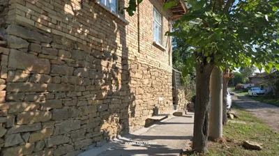 Brick house side with stone foundation, concrete walkway, pruned trees, dirt road with parked cars and greenery.