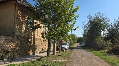 Brick house side with stone foundation, green door, pruned trees, utility pole, dirt road with parked cars and greenery.