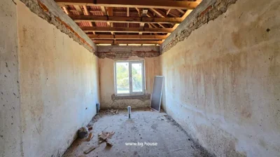 Room interior showing a window, exposed wooden roof beams, bare plaster walls, and a rough concrete floor with construction debris.