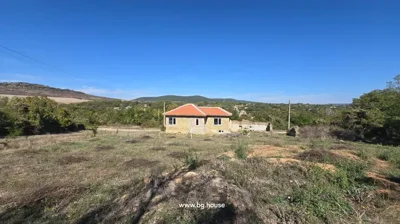 Brick house with red tiled roof surrounded by grass, trees, and hills in the distance.