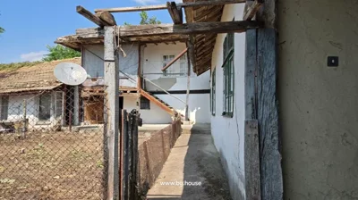 Narrow concrete pathway alongside a traditional Bulgarian house with wooden stairs, fenced yard, and visible windows under a clear sky.
