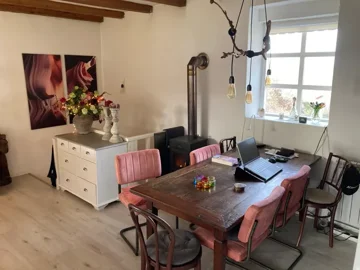 Dining area with a wood-burning stove, a white dresser with decorative items, wall art, and a wooden table with pink and wooden chairs.